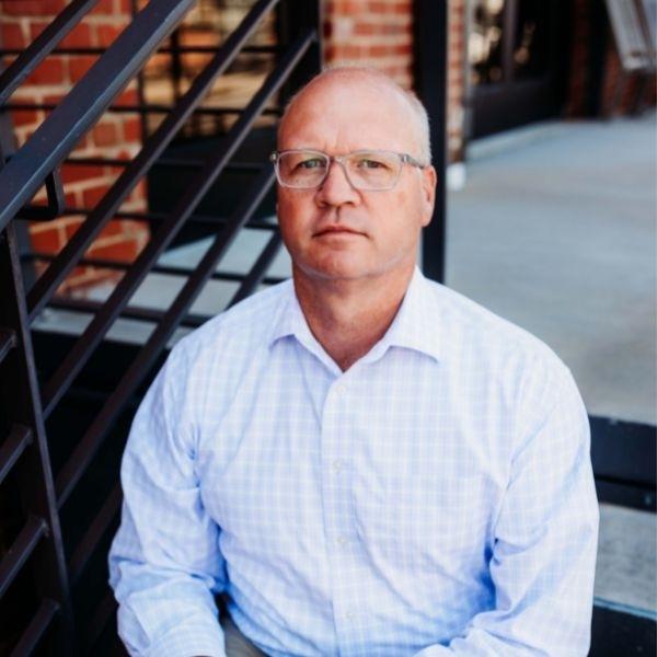 Dave Ingram wearing a light blue shirt and glasses sitting on some steps by a brick building